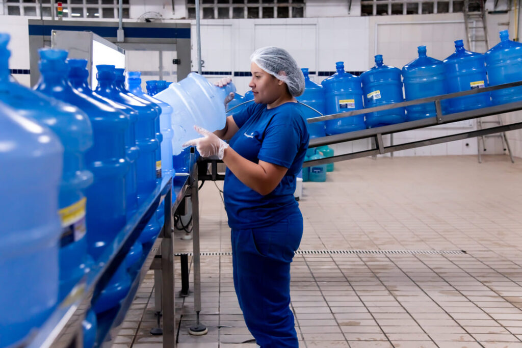 A imagem mostra uma funcionária em uma linha de produção de água mineral em garrafões de 20 litros. Ela está vestindo uniforme azul, com touca de proteção no cabelo, luvas e máscara facial. Ela está segurando um garrafão transparente, inspecionando-o cuidadosamente. O ambiente é limpo, com piso cerâmico claro e várias garrafas alinhadas sobre esteiras transportadoras.